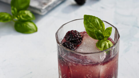 an old fashioned glass filled with a purple blackberry bourbon smash garnished with a large ice cube, blackberries, and basil