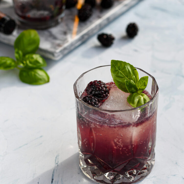 an old fashioned glass filled with a purple blackberry bourbon smash garnished with a large ice cube, blackberries, and basil