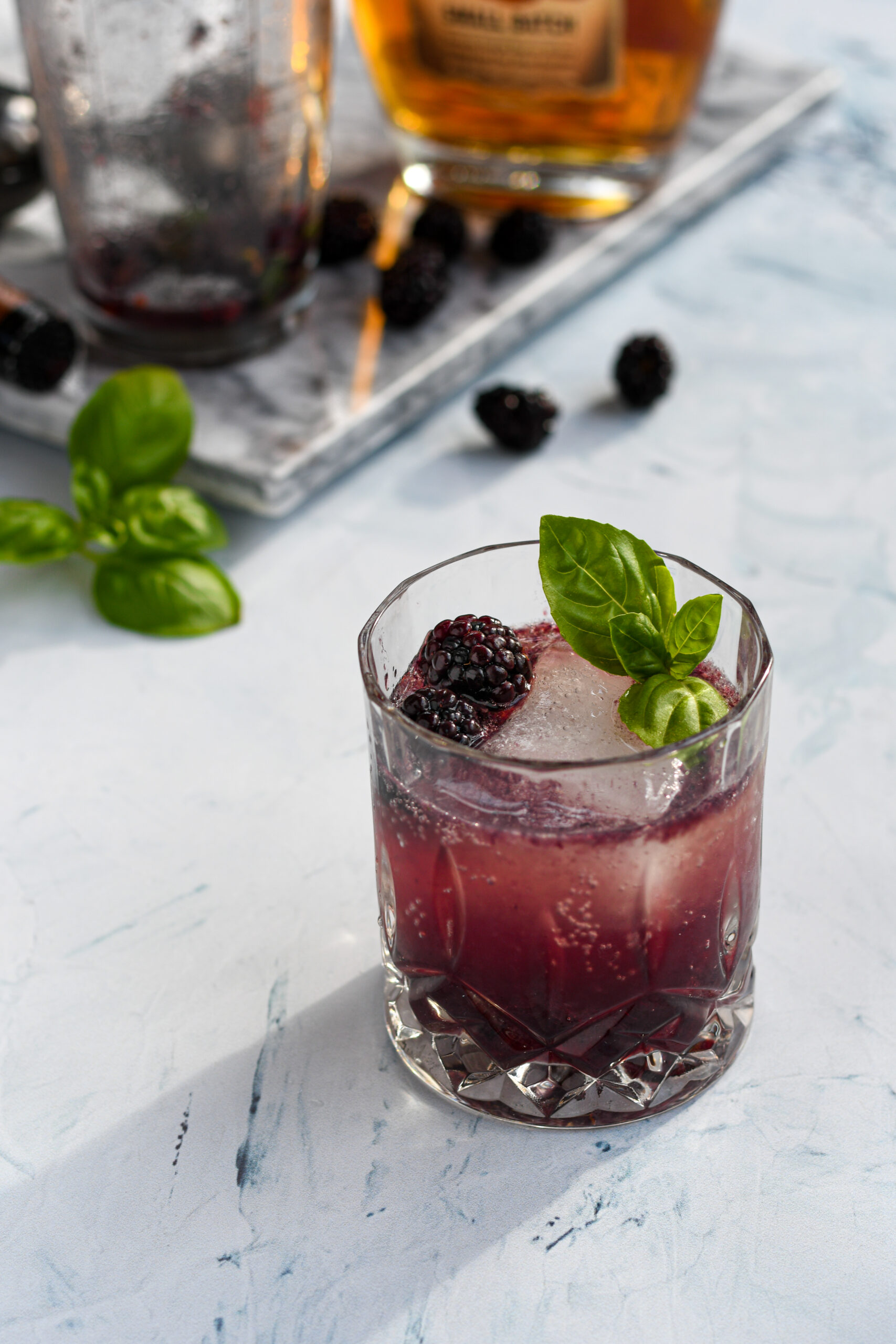 an old fashioned glass filled with a purple blackberry bourbon smash garnished with a large ice cube, blackberries, and basil