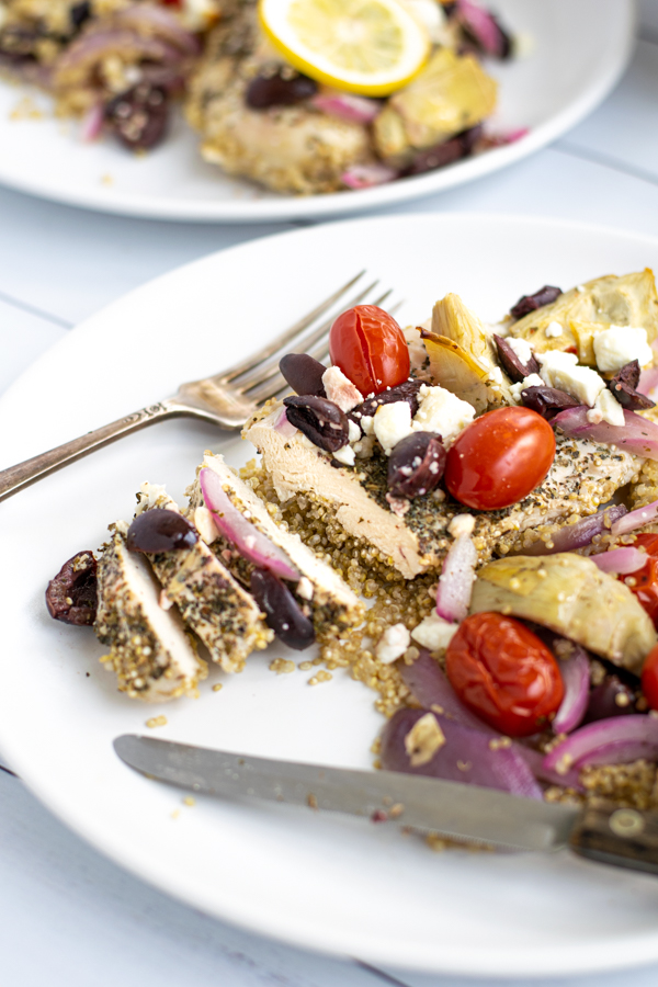 a cut chicken breast with greek toppings and quinoa on a white plate with a knife and fork
