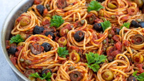 skillet on a table full of puttanesca pasta garnished with olives and parsley