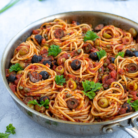 skillet on a table full of puttanesca pasta garnished with olives and parsley