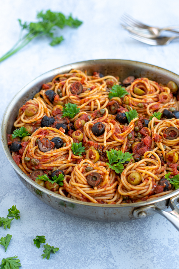 skillet on a table full of puttanesca pasta garnished with olives and parsley
