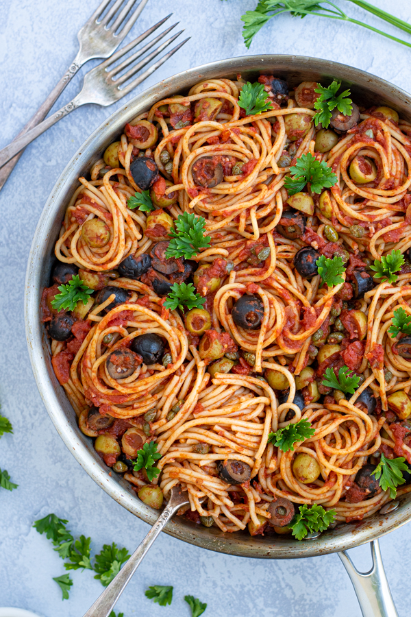 a fork spiraling some spaghetti puttanesca in a large skillet garnished by parsley