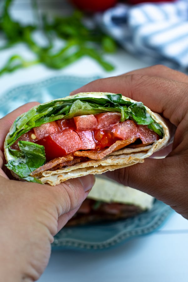 a cut half of a BLT wrap in a pair of hands showing off the bacon, lettuce, and tomato on the inside