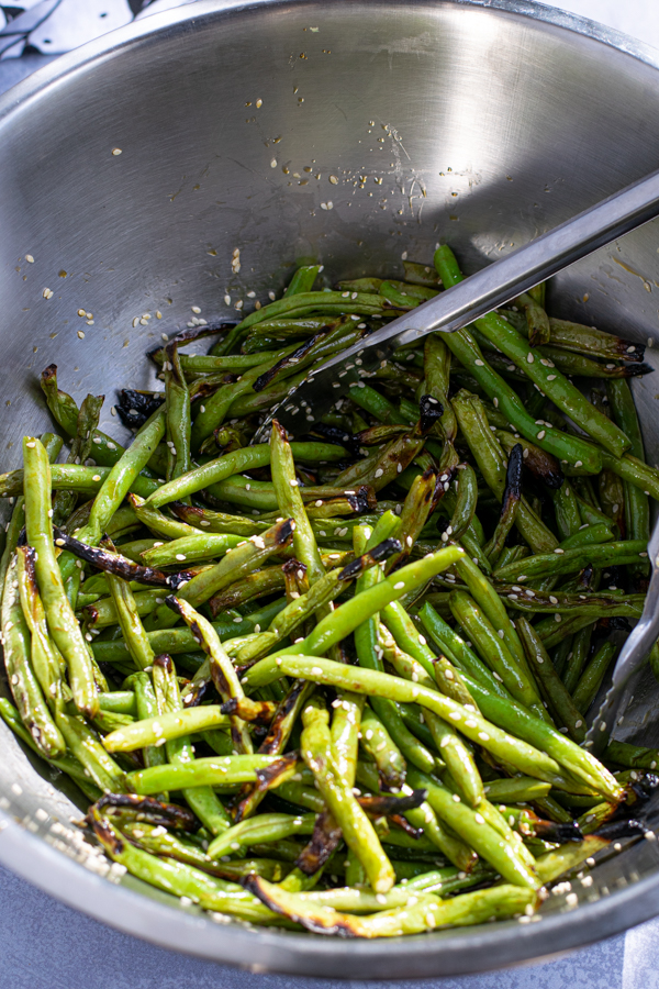 grilled green beans in a large bowl with tongs tossed in teriyaki sauce and sesame seeds