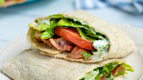 two halves of a BLT wrap on a white plate showing off the bacon, lettuce, tomato, and sauce on the inside