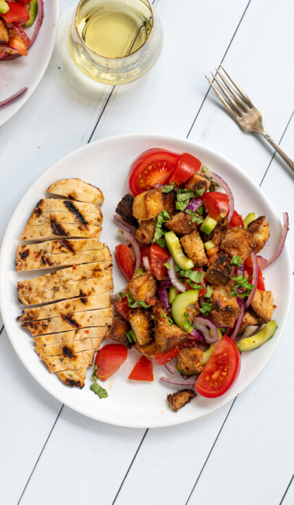 a white plate shot from overhead with sliced grilled chicken breast and a large serving of panzanella salad with tomatoes and grilled bread