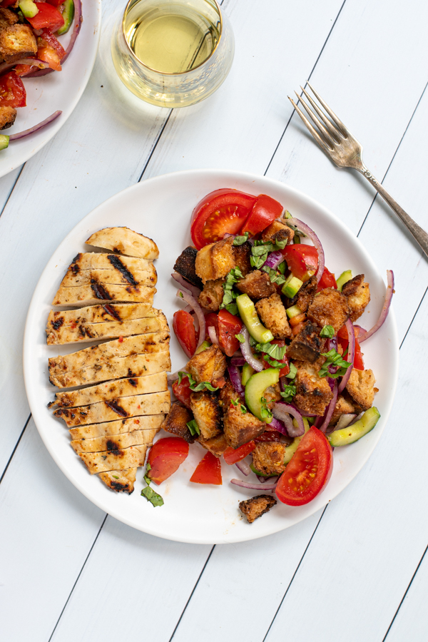 a white plate shot from overhead with sliced grilled chicken breast and a large serving of panzanella salad with tomatoes and grilled bread