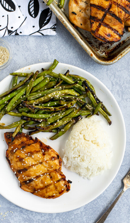 Overhead shot of a white plate with a teriyaki grilled chicken breast, grilled green beans with sauce, and white rice