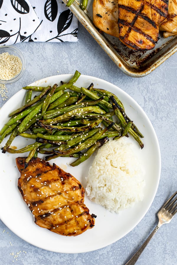 Overhead shot of a white plate with a teriyaki grilled chicken breast, grilled green beans with sauce, and white rice