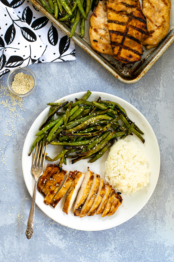 overhead view of a white plate filled with green beans, cut up chicken, and rice next to a serving platter with more grilled chicken