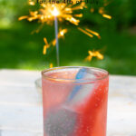 Pinterest Pin for red, white, and blue ice cubes showing a glass filled with red, white, and blue ice cubes on a table outside net to a sparkler