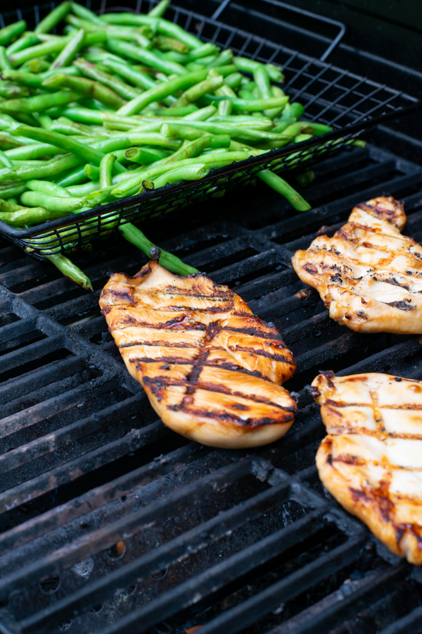 teriyaki marinated chicken breast on the grill next to a grill basket filled with fresh green beans