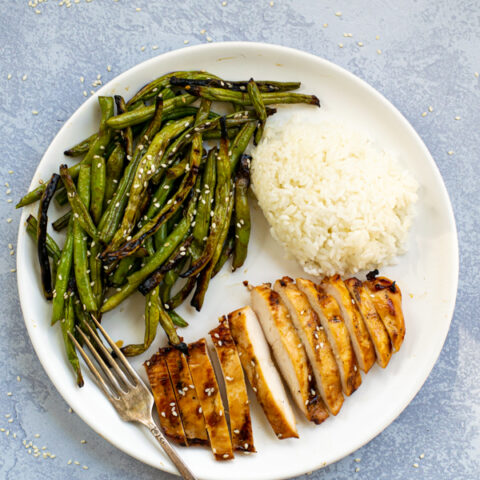 a white plate with teriyaki grilled chicken sliced up, grips grilled beans covered in sauce, and a pile of steamed white rice