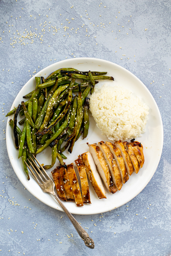 a white plate with teriyaki grilled chicken sliced up, grips grilled beans covered in sauce, and a pile of steamed white rice