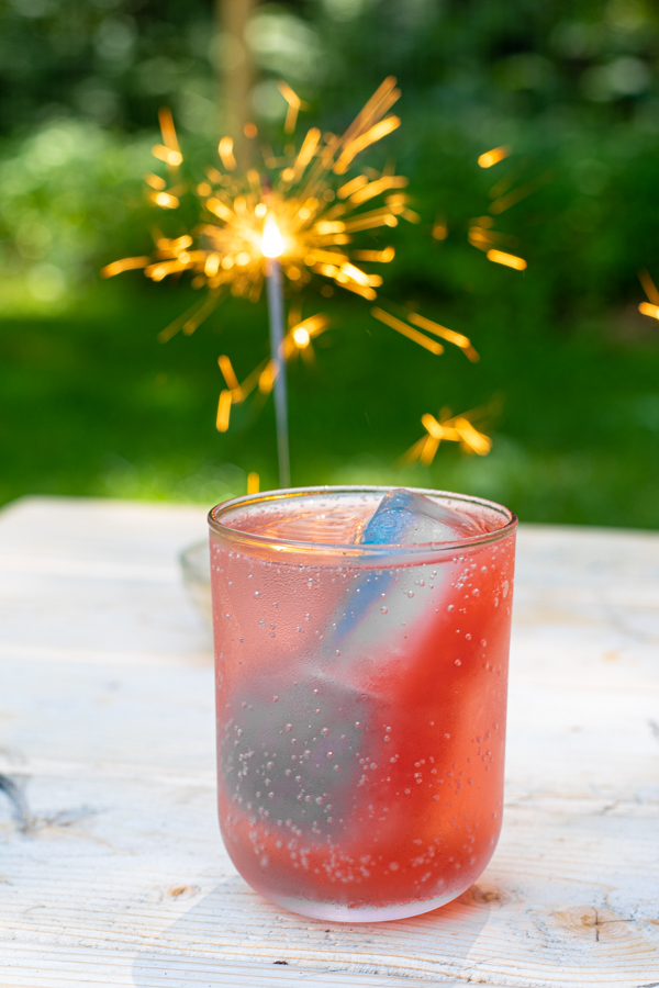 a glass filled with a clear beverage and layered red, white, and blue ice cubes on an outside table with a festive sparkler next to the glass