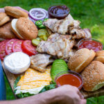 Pinterest Pin for Grilled Chicken Sandwich Board showing a man outside holding a large board filled with chicken sandwich toppings and chicken