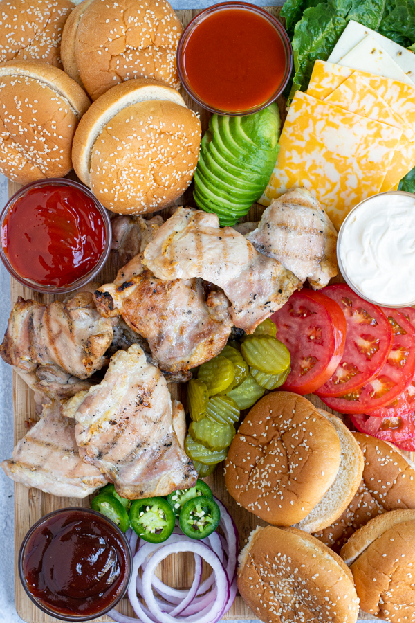close up of grilled chicken on a large bamboo cutting board with different sandwich toppings around the chicken