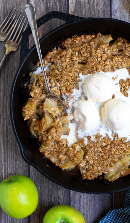 overhead shot of a cast iron on a table filled with apple crisp topped with ice cream and part of it scooped up with a spoon