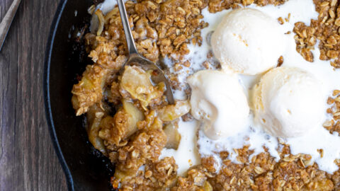 overhead shot of a cast iron on a table filled with apple crisp topped with ice cream and part of it scooped up with a spoon