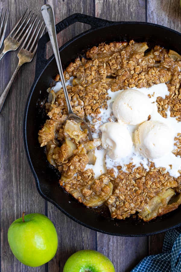 overhead shot of a cast iron on a table filled with apple crisp topped with ice cream and part of it scooped up with a spoon