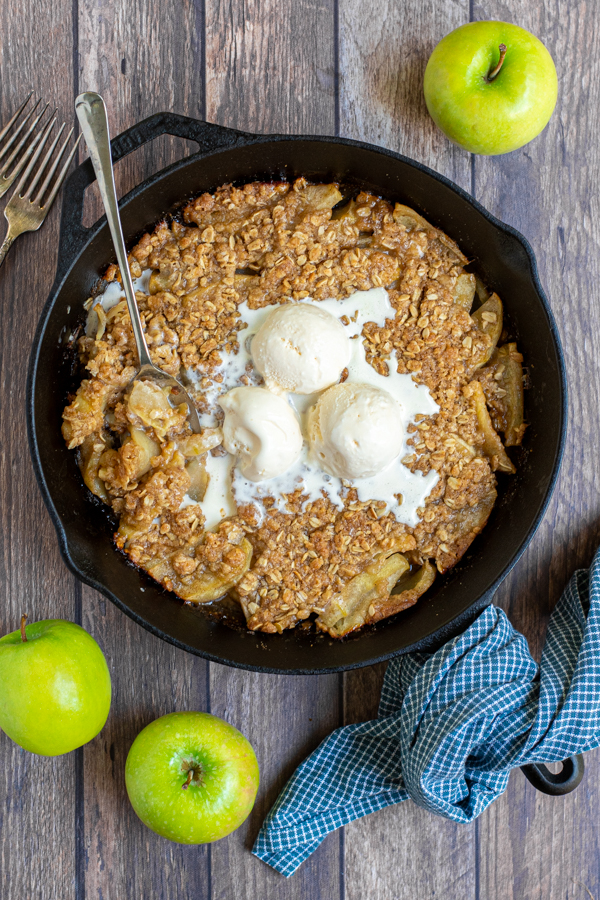 overhead view of a large cast iron pan filled with apple crisp topped with 3 scoops of ice cream and a serving spoon ready to dish out a scoop