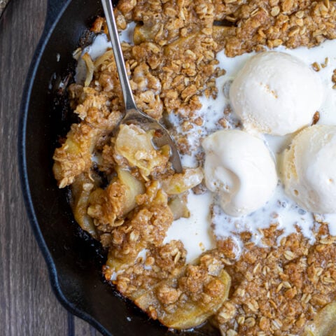 a serving spoon taking a scoop on apple crisp out of a pan and melted vanilla ice cream filling in the crisps empty scooped area