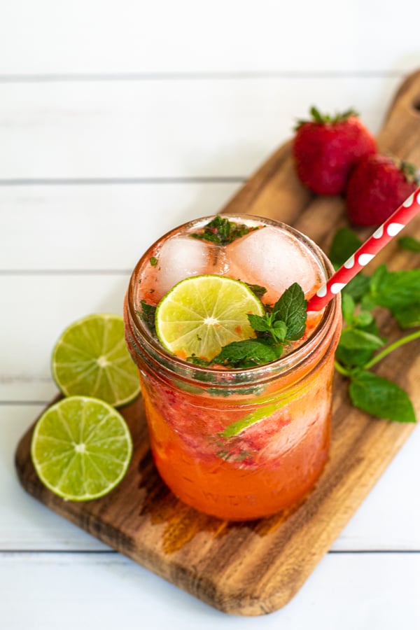 a glass of strawberry mojito mocktail on a wooden cutting board with cut limes and fresh mint leaves as garnishes