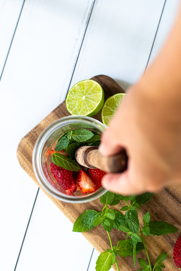 fresh cut strawberries and mint leaves at the bottom of a glass about to be muddled by a wooden muddler