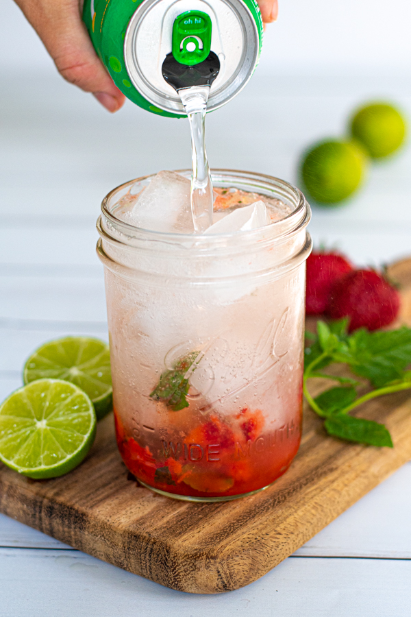 a lime seltzer being poured into a glass full of ice and muddled strawberries and mint