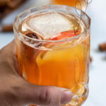 Pinterest Pin with text overlay Apple Cider Bourbon Cocktail, image of hand holding a whiskey glass full of bourbon cider garnished with cinnamon stick and apple.