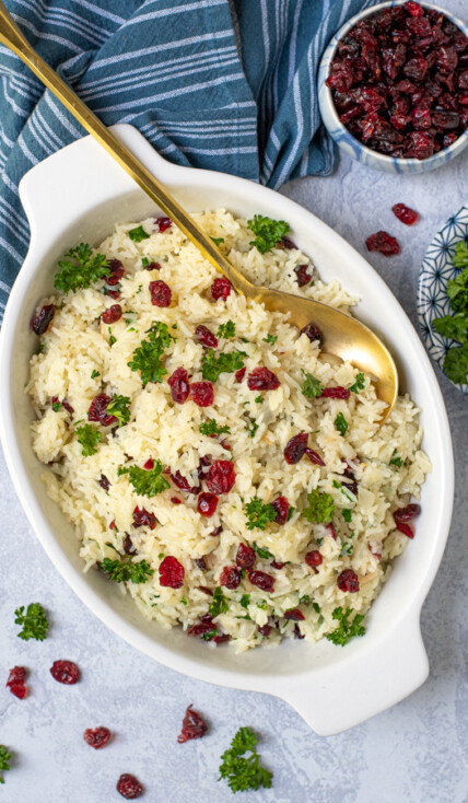 Rice Pilaf with Cranberries in a white oval serving dish with a matte gold serving spoon, garnished with dried cranberries and fresh parsley.