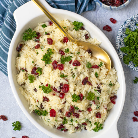 Rice Pilaf with Cranberries in a white oval serving dish with a matte gold serving spoon, garnished with dried cranberries and fresh parsley.