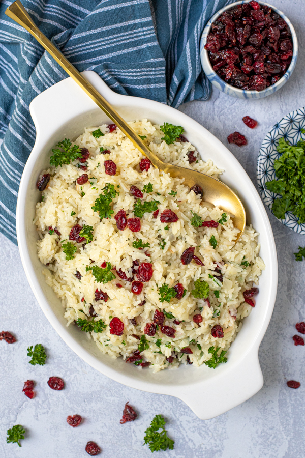Rice Pilaf with Cranberries in a white oval serving dish with a matte gold serving spoon, garnished with dried cranberries and fresh parsley.