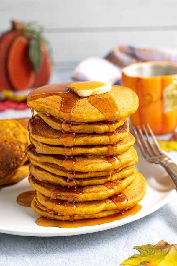 Stack of fluffy looking pumpkin pancakes on a white plate topped with a pad of butter and maple syrup running down the sides and pooling on the plate.