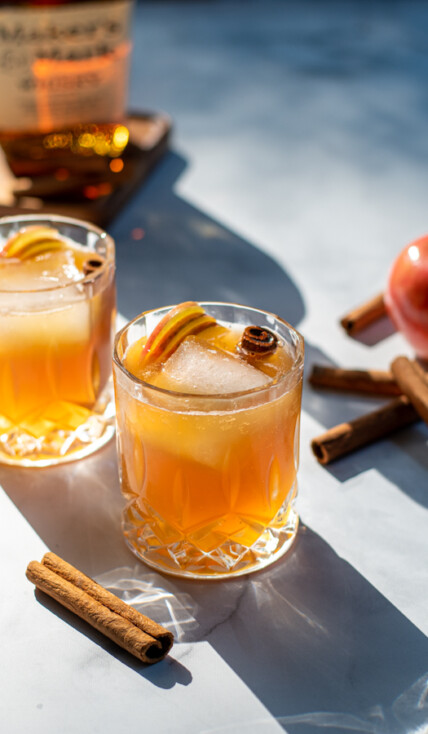 Hard sun light hitting two nice whiskey glasses full of apple cider bourbon cocktail garnished with cinnamon sticks and apple slices.