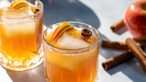 Hard sun light hitting two nice whiskey glasses full of apple cider bourbon cocktail garnished with cinnamon sticks and apple slices.