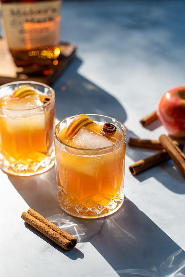 Hard sun light hitting two nice whiskey glasses full of apple cider bourbon cocktail garnished with cinnamon sticks and apple slices.