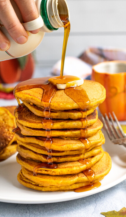 A tall stack of pumpkin pancakes on a white plate, a hand is pouring maple syrup from a jug over the top of the pancakes leaving the syrup to drip down the sides of the stack.