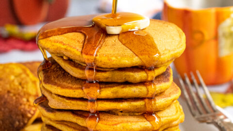 A tall stack of pumpkin pancakes on a white plate, a hand is pouring maple syrup from a jug over the top of the pancakes leaving the syrup to drip down the sides of the stack.