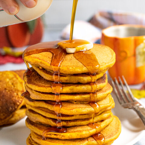 A tall stack of pumpkin pancakes on a white plate, a hand is pouring maple syrup from a jug over the top of the pancakes leaving the syrup to drip down the sides of the stack.