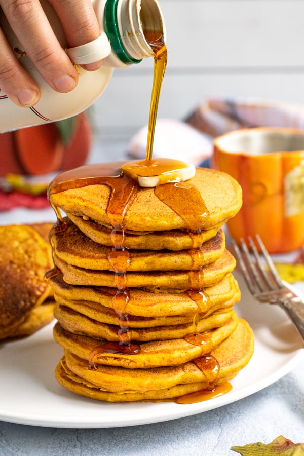 A tall stack of pumpkin pancakes on a white plate, a hand is pouring maple syrup from a jug over the top of the pancakes leaving the syrup to drip down the sides of the stack.