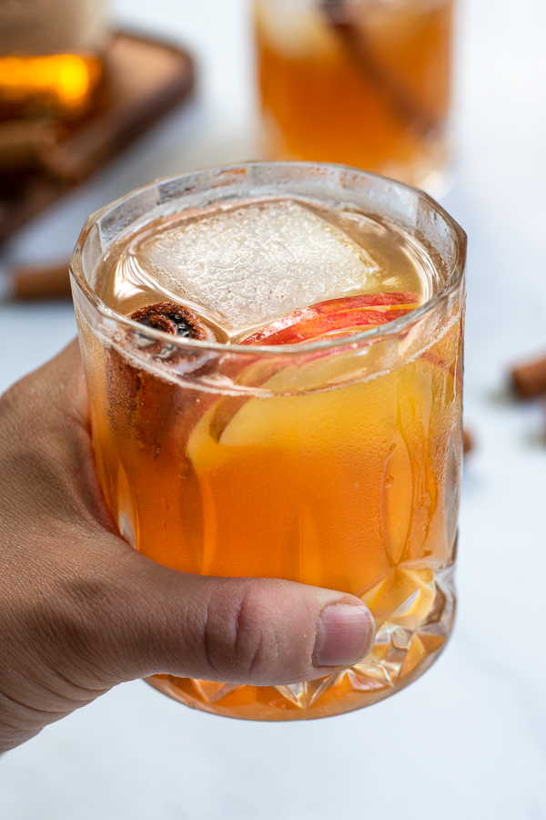 A hand holding a fancy whiskey glass full of Bourbon Apple Cider with a large ice cube, cinnamon stick, and apple slices.