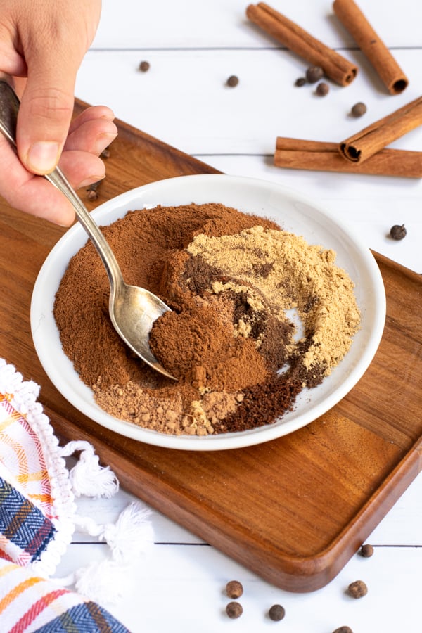 On a small white plate a spoon is stirring together the piles of different spices used to make pumpkin pie spice mix.