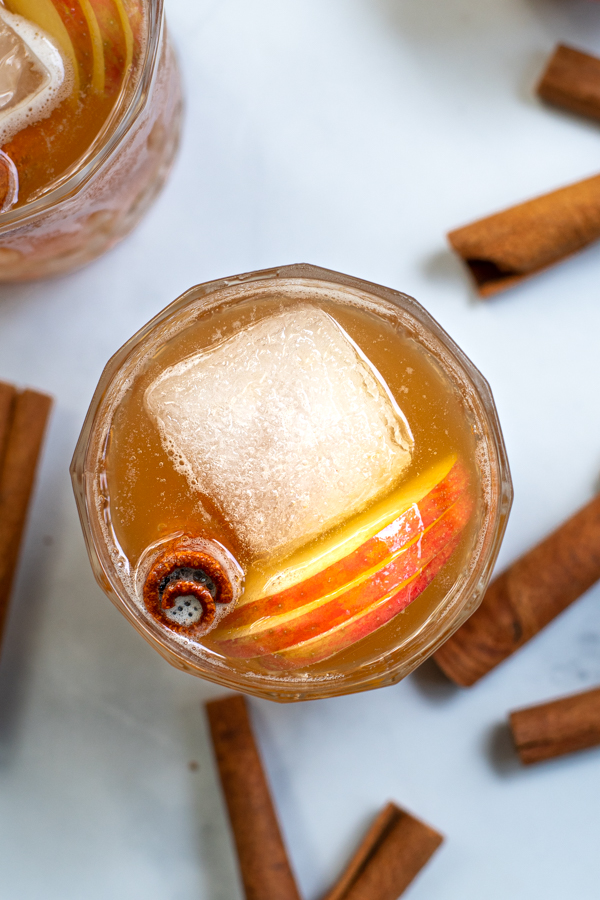 Overhead close up of an apple cider bourbon cocktail in a whiskey glass with a large ice cube, cinnamon stick, and thin apple slices.