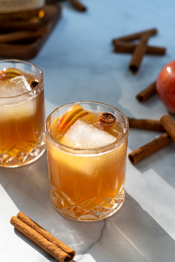 Two whiskey glasses full of apple cider bourbon garnished with a cinnamon stick and apple slices.