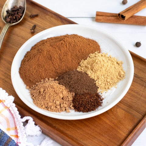 a small white plate sit-in non a wood board filled with 5 different spices used to make pumpkin spice.
