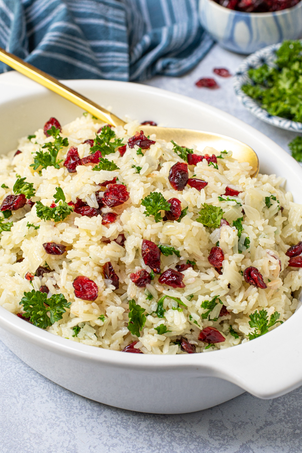 A white serving dish with a gold spoon in white rice pilaf with dried cranberries and fresh parsley.