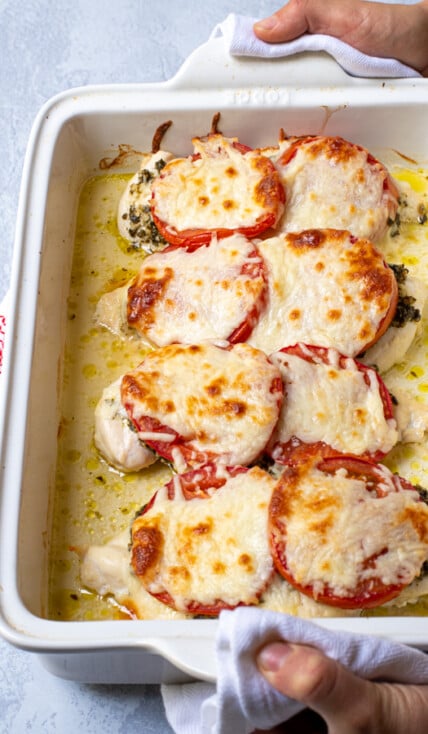 Hands holding a white baking dish containing pesto chicken bake, showing off melted mozzarella cheese on top.